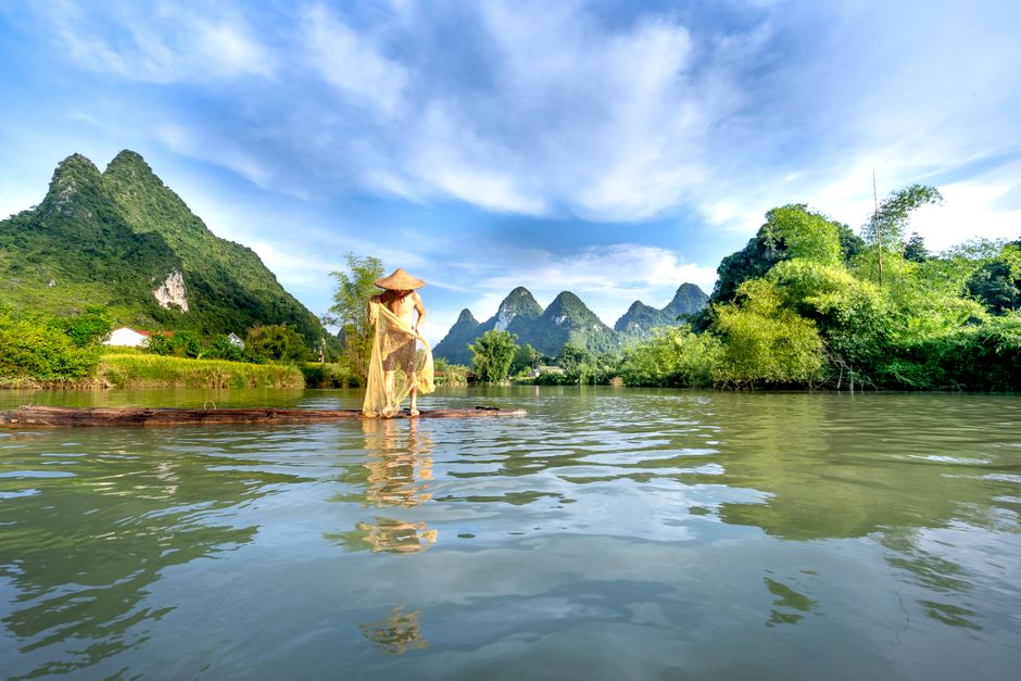 A person in a conical hat stands by a tranquil river with lush mountains in the background