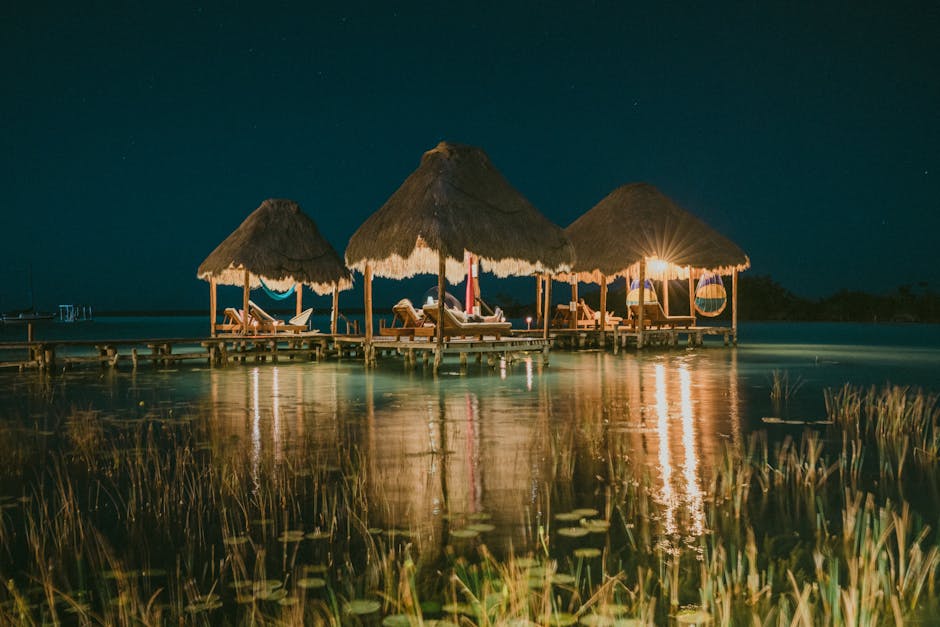 Illuminated tropical gazebos with hammocks reflect on a serene lake at night, creating a tranquil resort scene.