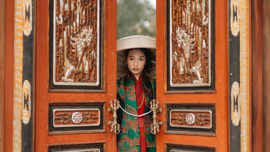 A woman in traditional Vietnamese attire stands at a ornate temple door in Hanoi, Vietnam.
