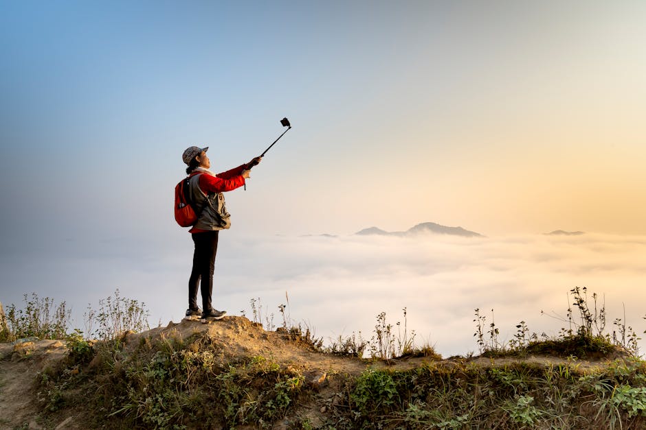 Young woman takes a selfie on a mountain peak at sunrise, surrounded by clouds and nature.
