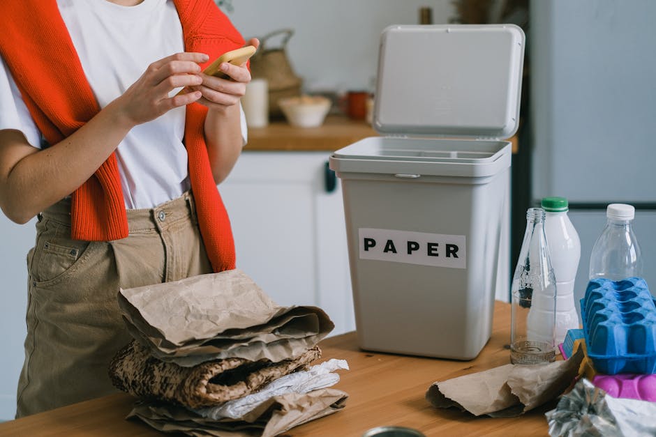 Crop unrecognizable person using smartphone while sorting waste on wooden table on blurred background