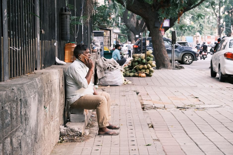 An adult man sitting on a sidewalk, engaged in a phone call in Bengaluru, India.