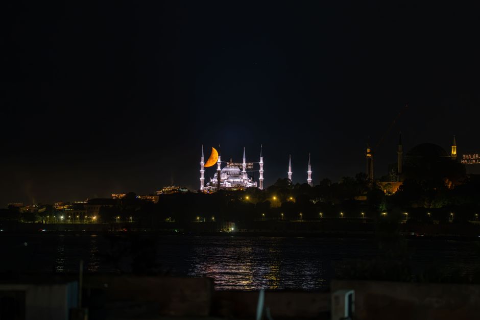 Majestic night view of the Blue Mosque in Istanbul with a crescent moon and city lights reflecting on the water.