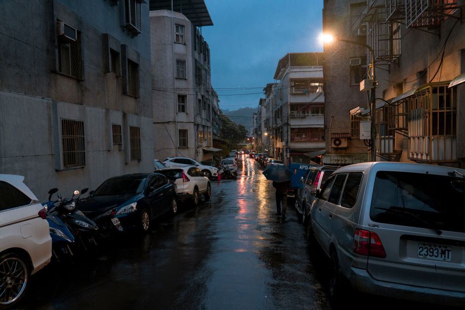 Moody urban street during a rainy night with reflections, parked cars, and streetlights.