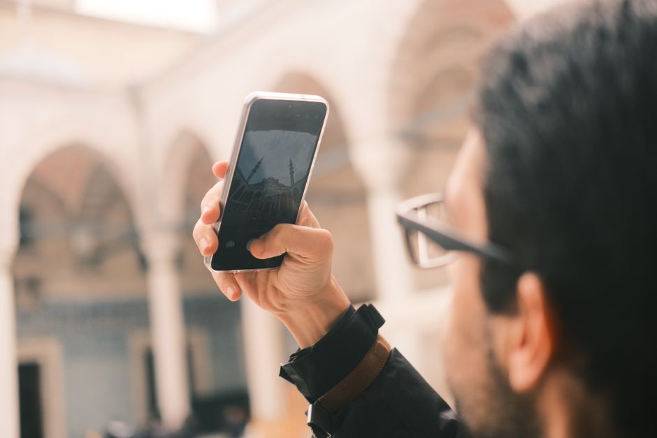 An adult male photographs a mosque using a smartphone outdoors with a focus on capturing architecture.