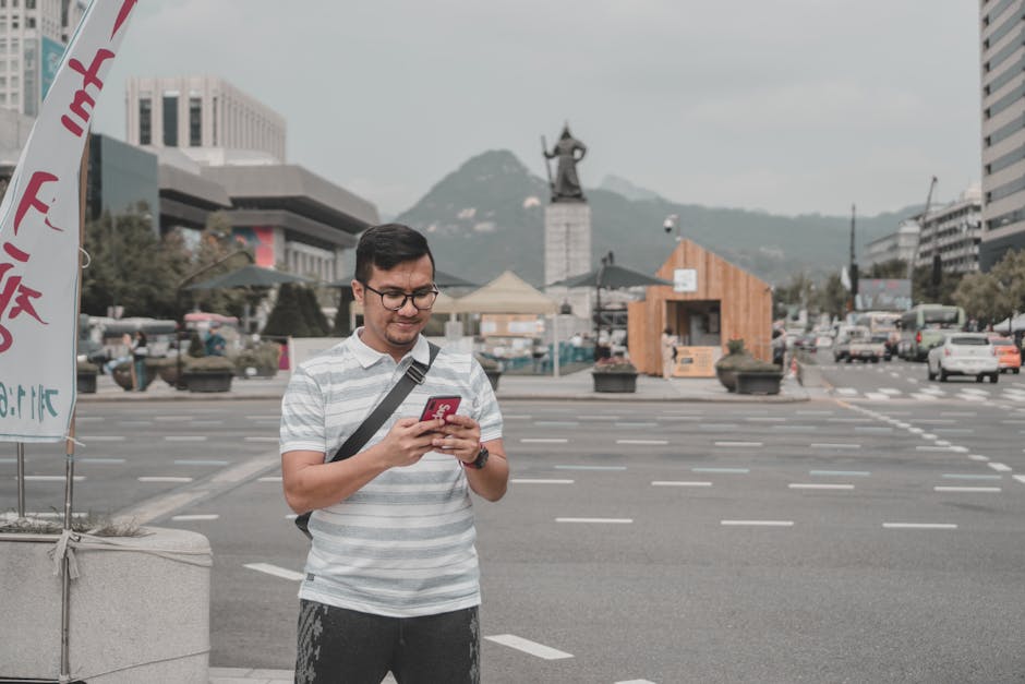 A man in a striped shirt standing on a street using a smartphone with a city skyline in the background.