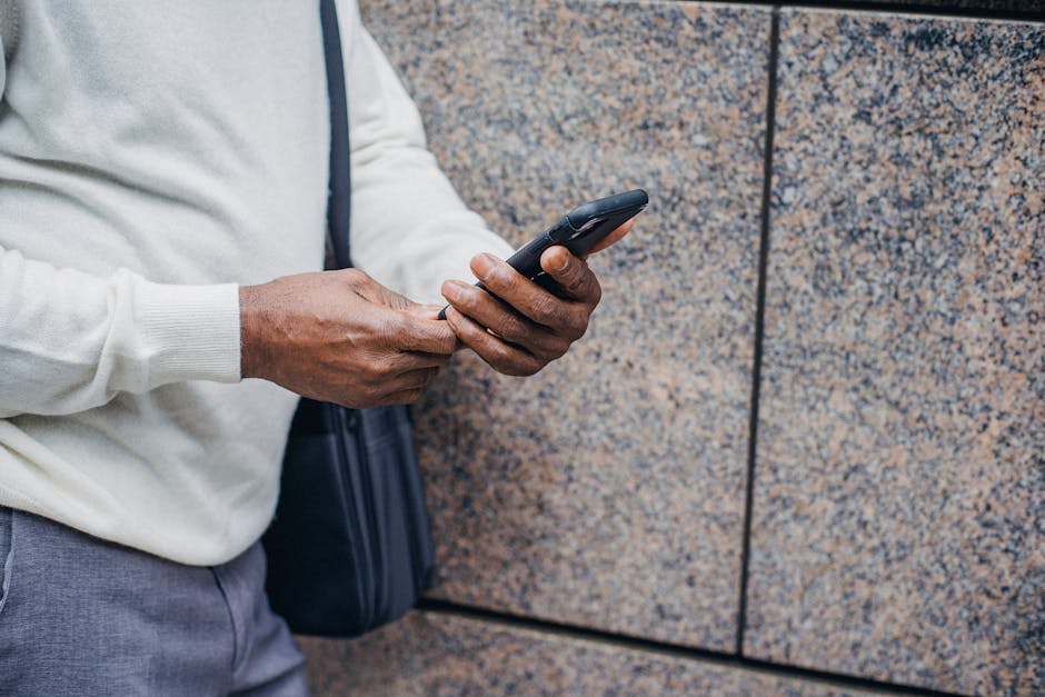 Crop faceless black businessman in trendy outfit browsing smartphone while leaning on wall